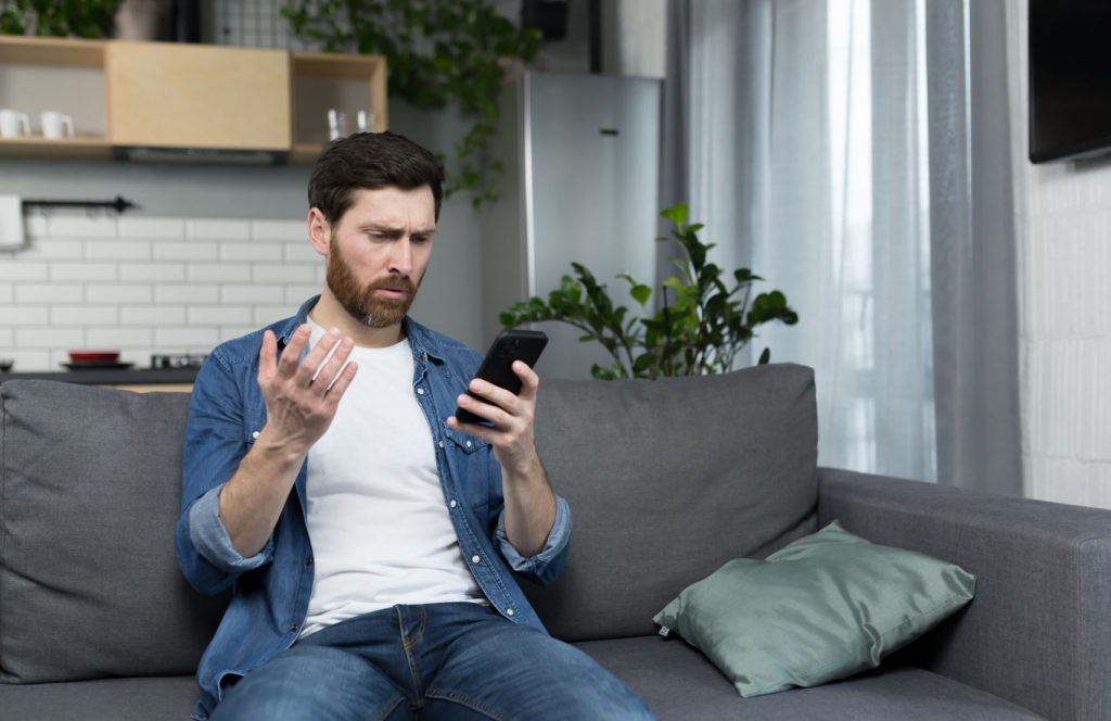 Homem frustrado sentado em sofá olhando telefone