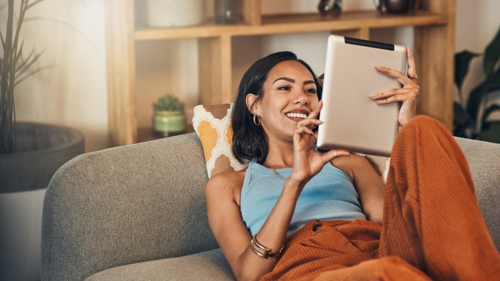 Mulher deitada em sofá sorrindo e assistindo tablet